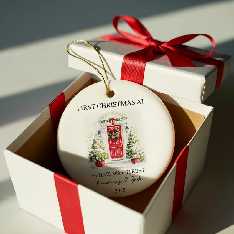 Puede incluir: Un adorno navide&ntilde;o de cer&aacute;mica blanca con una cuerda dorada, dentro de una caja de regalo blanca con una cinta roja. El adorno presenta una ilustraci&oacute;n de acuarela de una puerta principal roja con una corona, el texto "FIRST CHRISTMAS AT", direcci&oacute;n y nombres.