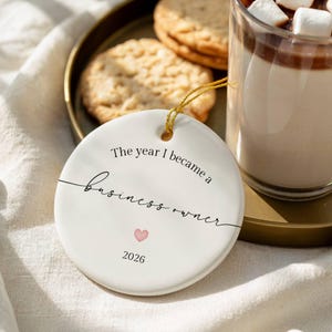 May include: A round white ceramic ornament featuring black text that reads The year I became a business owner, a small pink heart, and the year 2026. The ornament has a gold string loop at the top for hanging and a smooth, glossy finish.