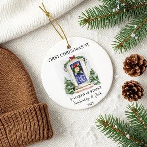 Puede incluir: Adorno navide&ntilde;o de cer&aacute;mica blanca con una cuerda dorada. El adorno presenta una ilustraci&oacute;n en acuarela de una puerta azul con una corona, el texto "FIRST CHRISTMAS AT", direcci&oacute;n, nombres y el a&ntilde;o 2025.