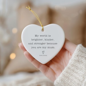 Peut inclure: Ornement blanc en forme de cœur avec une ficelle dorée, tenu dans une main. L'ornement porte le texte "My world is brighter, kinder, and stronger because you are my mum." et "Love Cindy" sous un symbole de cœur.