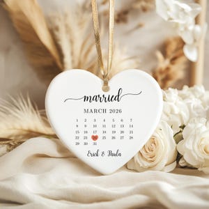 May include: A white heart-shaped ornament with the word "married" and a calendar for March 2026. The date 24 is highlighted in red. The names "Erick & Paula" are at the bottom. A gold ribbon is attached for hanging.