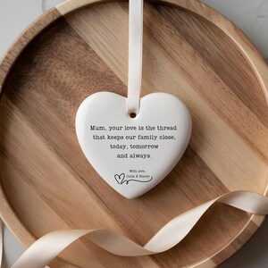 Pode incluir: Um ornamento de cerâmica branca em forma de coração pendurado por uma fita cor de creme. O texto diz: Mum, your love is the thread that keeps our family close, today, tomorrow and always. With love, Julia & Stacey, com um pequeno gráfico de coração.