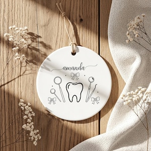 Può includere: Ornamento a disco in ceramica bianca a tema dentale. Presenta un dente, strumenti odontoiatrici e il nome "amanda" in corsivo. Il design è in nero, con un cordino per appenderlo. Sfondo in legno.