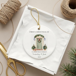 Puede incluir: Adorno navide&ntilde;o redondo de cer&aacute;mica blanca con una anilla dorada. Presenta una ilustraci&oacute;n en acuarela de una puerta principal con una corona y el texto "FIRST CHRISTMAS AT". La direcci&oacute;n "53 HARTWAY STREET", nombres y el a&ntilde;o 2025 est&aacute;n debajo.