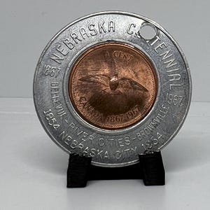 May include: A silver-colored commemorative coin with a hole in the top. The coin features the text "Nebraska Centennial 1867-1967" around the edge. In the center is a copper-colored coin with the text "1 Cent" and a bird in flight. The text "Canada 1867-1967" is also visible on the copper coin.