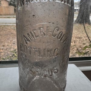 May include: A clear glass bottle with embossed text. The bottle reads "REGISTERED" at the top and "AMERI-COLA BOTTLING CO. AMERICUS GA." in a circular design. The bottle is vintage and shows signs of age.