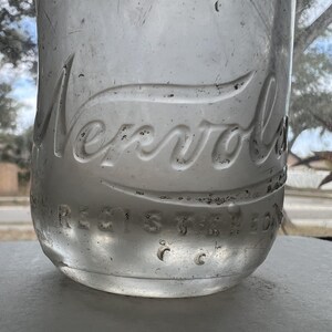 May include: Close-up of a clear glass bottle with the embossed word "Nervol" in a curved script. The word "REGISTERED" is also embossed on the bottle. The bottle is sitting on a white surface.
