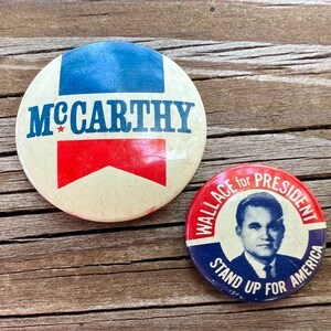 May include: Two vintage political campaign buttons. The first button is white with blue and red stripes and the text "McCarthy" in blue. The second button is red, white, and blue with the text "Wallace for President Stand Up For America".