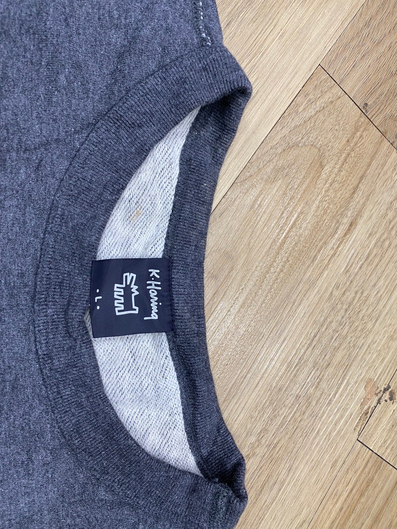 May include: Dark gray crew neck sweatshirt with a white interior. The label reads "K. Haring" with a stylized graphic and the size "-L."