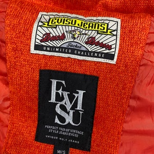 May include: A close-up of a bright orange jacket with a black EVISU JEANS label. The label has the text "Duck Down Jacket Unlimited Challenge" and "Perfect Pair of Vintage Style Jeans EVG.52 Unique No.1 Jeans W/S".