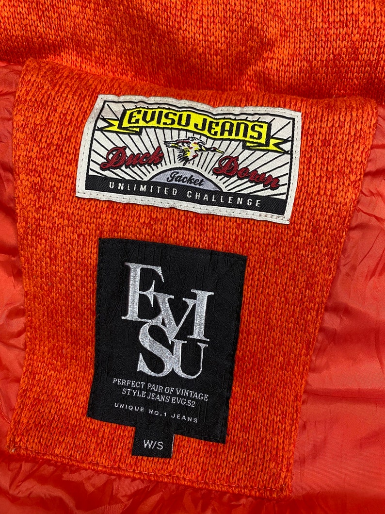 May include: A close-up of a bright orange jacket with a black EVISU JEANS label. The label has the text "Duck Down Jacket Unlimited Challenge" and "Perfect Pair of Vintage Style Jeans EVG.52 Unique No.1 Jeans W/S".
