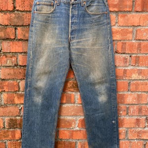 May include: Vintage blue denim jeans with a button fly, classic five-pocket design, and faded areas. The jeans are hanging on a gold-colored hanger against a red brick wall. The jeans appear to be a straight-leg style.