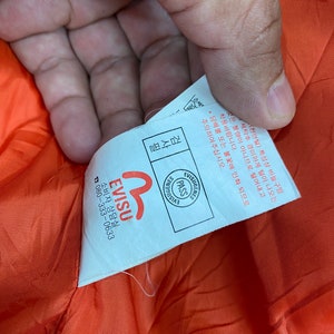 May include: White tag with black text that reads "Evisu" and a red logo. The tag also has a smaller white tag with black text that reads "Visugenies Pass Evisu". The tag is attached to a bright orange fabric.