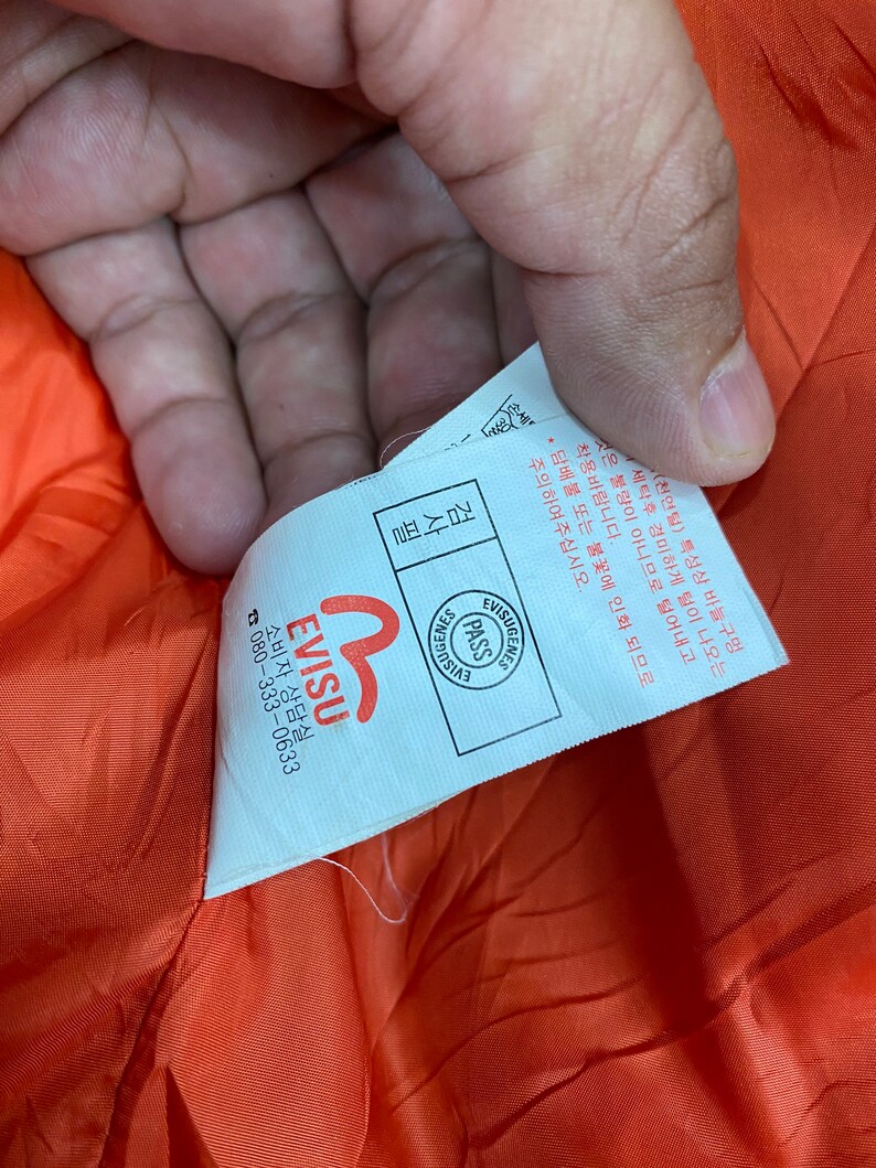 May include: White tag with black text that reads "Evisu" and a red logo. The tag also has a smaller white tag with black text that reads "Visugenies Pass Evisu". The tag is attached to a bright orange fabric.