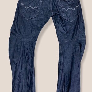 Peut inclure: Un jean en denim bleu fonc&eacute; avec plusieurs poches et des d&eacute;tails de couture. Le jean a une coupe d&eacute;contract&eacute;e et une jambe droite.