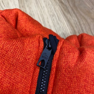 May include: Close-up of a black zipper with a black pull tab that says "EVISU" on an orange knitted fabric.