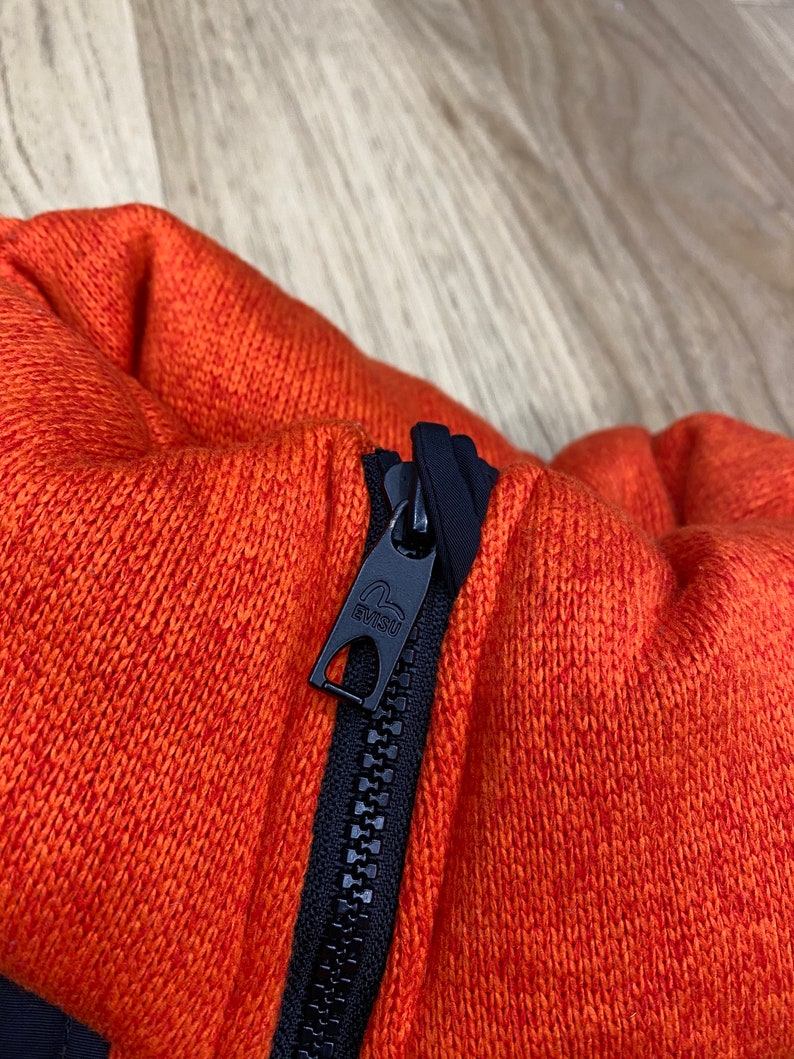 May include: Close-up of a black zipper with a black pull tab that says "EVISU" on an orange knitted fabric.
