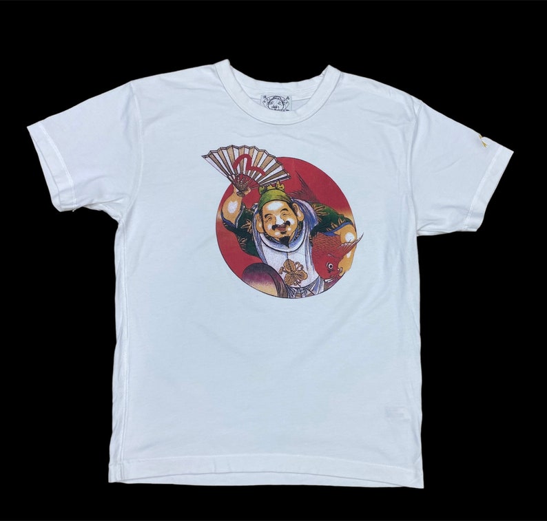 May include: White t-shirt with a red circle graphic featuring a cartoon character holding a fan. The character is wearing a yellow hat and a blue and red robe. The text "C" is visible on the graphic.