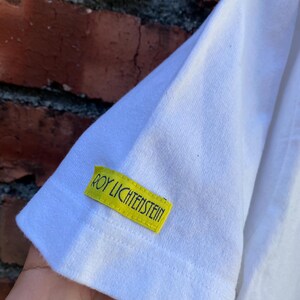 May include: White t-shirt sleeve with a yellow tag that reads "ROY LICHTENSTEIN" in green text. The tag is rectangular and sewn onto the sleeve. The background is a weathered brick wall.