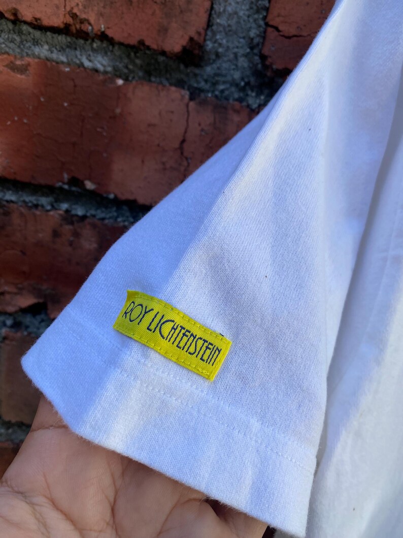 May include: White t-shirt sleeve with a yellow tag that reads "ROY LICHTENSTEIN" in green text. The tag is rectangular and sewn onto the sleeve. The background is a weathered brick wall.