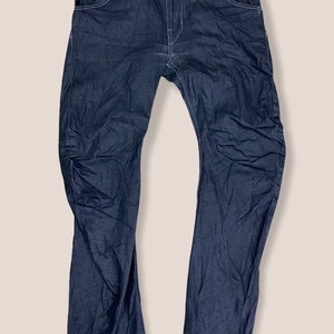Peut inclure: Un jean en denim bleu fonc&eacute; avec un petit patch de couleur or sur la poche avant gauche. Le patch porte le texte "G-Star RAW".