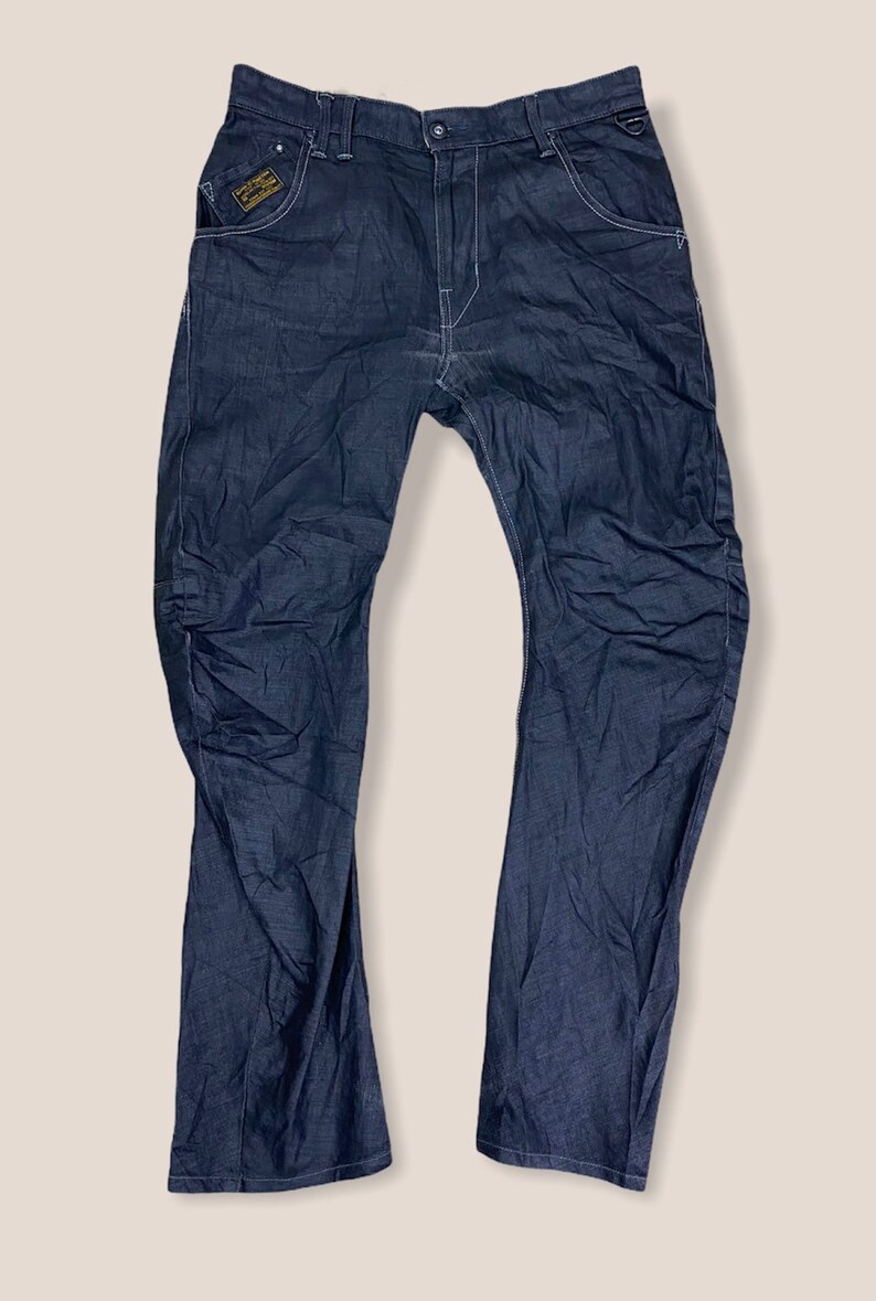 Peut inclure: Un jean en denim bleu fonc&eacute; avec un petit patch de couleur or sur la poche avant gauche. Le patch porte le texte "G-Star RAW".