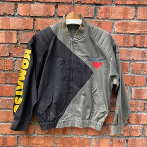 May include: A black and green bomber jacket with a yellow "KOMWISO" graphic on the left sleeve. The jacket has a zipper closure and a red triangle on the right side.