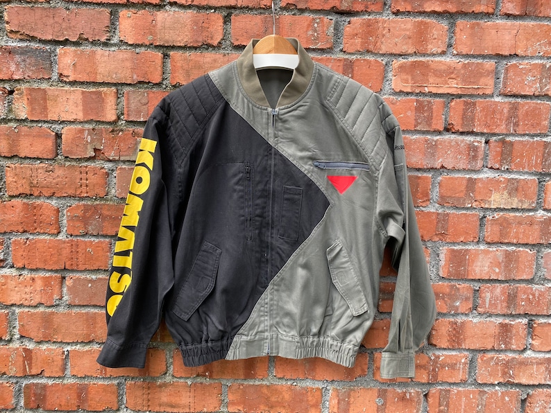May include: A black and green bomber jacket with a yellow "KOMWISO" graphic on the left sleeve. The jacket has a zipper closure and a red triangle on the right side.