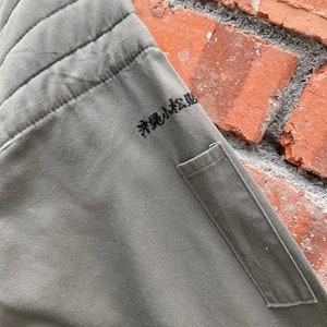 May include: A green quilted jacket with a small pocket on the left side. The jacket has Japanese text embroidered on the right side that reads "粋なは風"