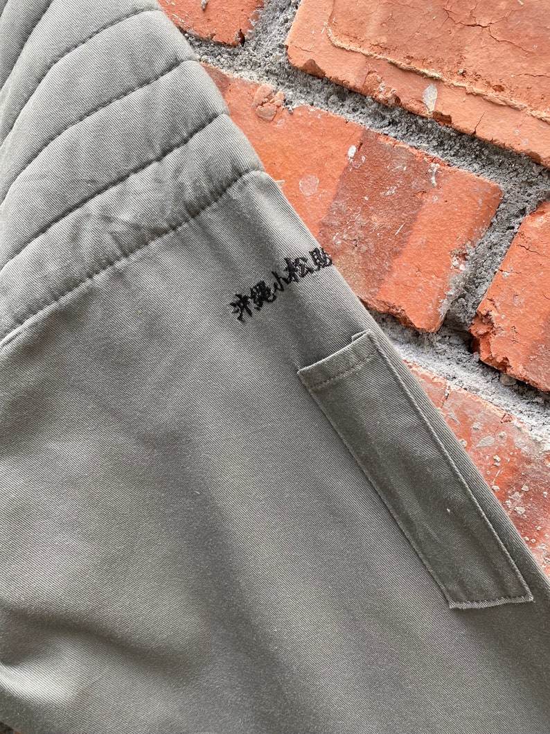 May include: A green quilted jacket with a small pocket on the left side. The jacket has Japanese text embroidered on the right side that reads "粋なは風"