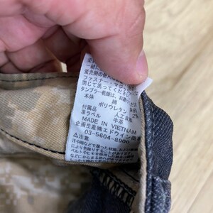 Peut inclure: Gros plan sur une &eacute;tiquette d'entretien avec des instructions de lavage et de s&eacute;chage. L'&eacute;tiquette est fix&eacute;e &agrave; un pantalon en jean noir avec un motif camouflage. L'&eacute;tiquette indique : "Ne pas utiliser avec de l'eau de Javel fluorescente. Fermer la fermeture &eacute;clair et la bande magique avant le lavage. Le s&eacute;chage en tambour est interdit. Corps principal : Coton, Accessoires : Polyur&eacute;thane 98%, &Eacute;tiquette en cuir : 2%, Cuir artificiel : Cuir de vache. Fabriqu&eacute; au Vietnam. Con&ccedil;u et produit par Edwin Trading Co., Ltd. 03-5604-8900. Attention : La sueur ou la pluie peuvent provoquer des d&eacute;colorations. Lorsque vous utilisez une ceinture ou une ficelle, faites attention aux enchev&ecirc;trements. Veuillez le porter serr&eacute;."