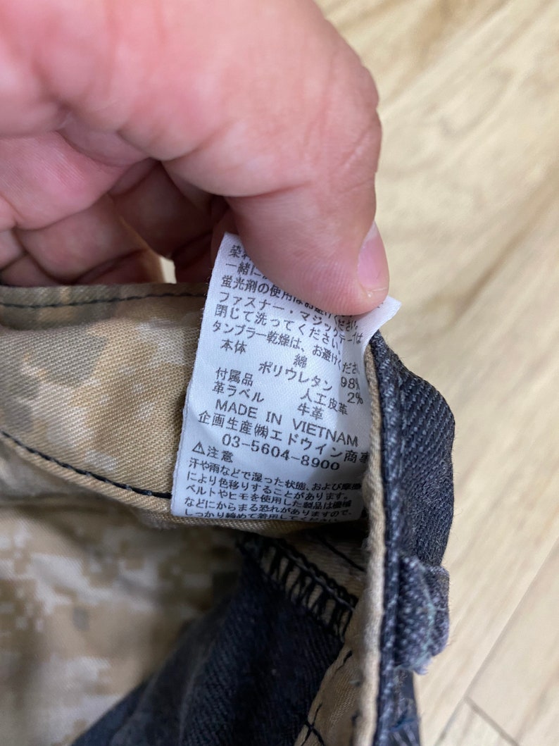 Peut inclure: Gros plan sur une &eacute;tiquette d'entretien avec des instructions de lavage et de s&eacute;chage. L'&eacute;tiquette est fix&eacute;e &agrave; un pantalon en jean noir avec un motif camouflage. L'&eacute;tiquette indique : "Ne pas utiliser avec de l'eau de Javel fluorescente. Fermer la fermeture &eacute;clair et la bande magique avant le lavage. Le s&eacute;chage en tambour est interdit. Corps principal : Coton, Accessoires : Polyur&eacute;thane 98%, &Eacute;tiquette en cuir : 2%, Cuir artificiel : Cuir de vache. Fabriqu&eacute; au Vietnam. Con&ccedil;u et produit par Edwin Trading Co., Ltd. 03-5604-8900. Attention : La sueur ou la pluie peuvent provoquer des d&eacute;colorations. Lorsque vous utilisez une ceinture ou une ficelle, faites attention aux enchev&ecirc;trements. Veuillez le porter serr&eacute;."