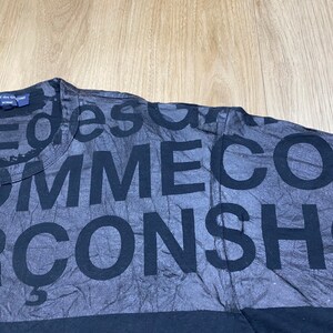 May include: Black t-shirt with a faded print of the words "Comme des Gar&ccedil;ons" repeated in a random pattern.