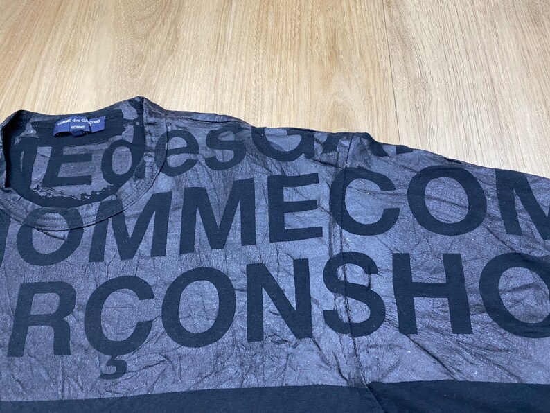May include: Black t-shirt with a faded print of the words "Comme des Gar&ccedil;ons" repeated in a random pattern.