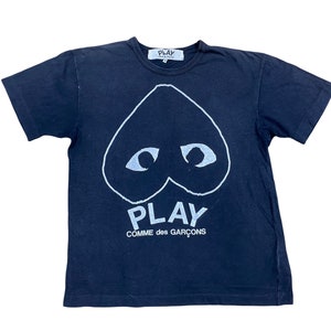 May include: Navy blue t-shirt featuring a white heart-shaped graphic with eyes and the word "PLAY" above "COMME des GARÇONS". The shirt has a crew neck and short sleeves. A small white tag is visible at the neckline.