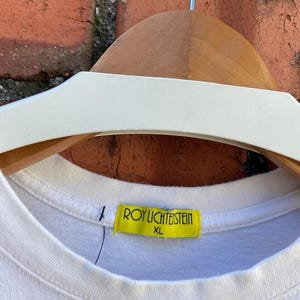 May include: White XL t-shirt with a crew neck, hanging on a wooden hanger. A yellow tag with the text "ROY LICHTENSTEIN" and "XL" is attached to the shirt's collar. The background features a brick wall.