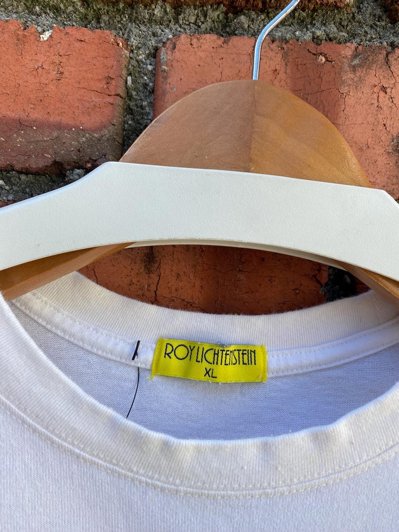 May include: White XL t-shirt with a crew neck, hanging on a wooden hanger. A yellow tag with the text "ROY LICHTENSTEIN" and "XL" is attached to the shirt's collar. The background features a brick wall.