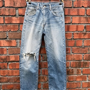 May include: Light blue denim jeans featuring a distressed finish with a prominent horizontal rip on the right knee, a classic five-pocket design, a metal button fly closure, and a straight-leg cut, displayed on a wooden hanger.
