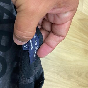 May include: A black t-shirt with a graphic print and a blue tag that reads "COMME des GARCONS*" and "HOMME" with a size "M" label.