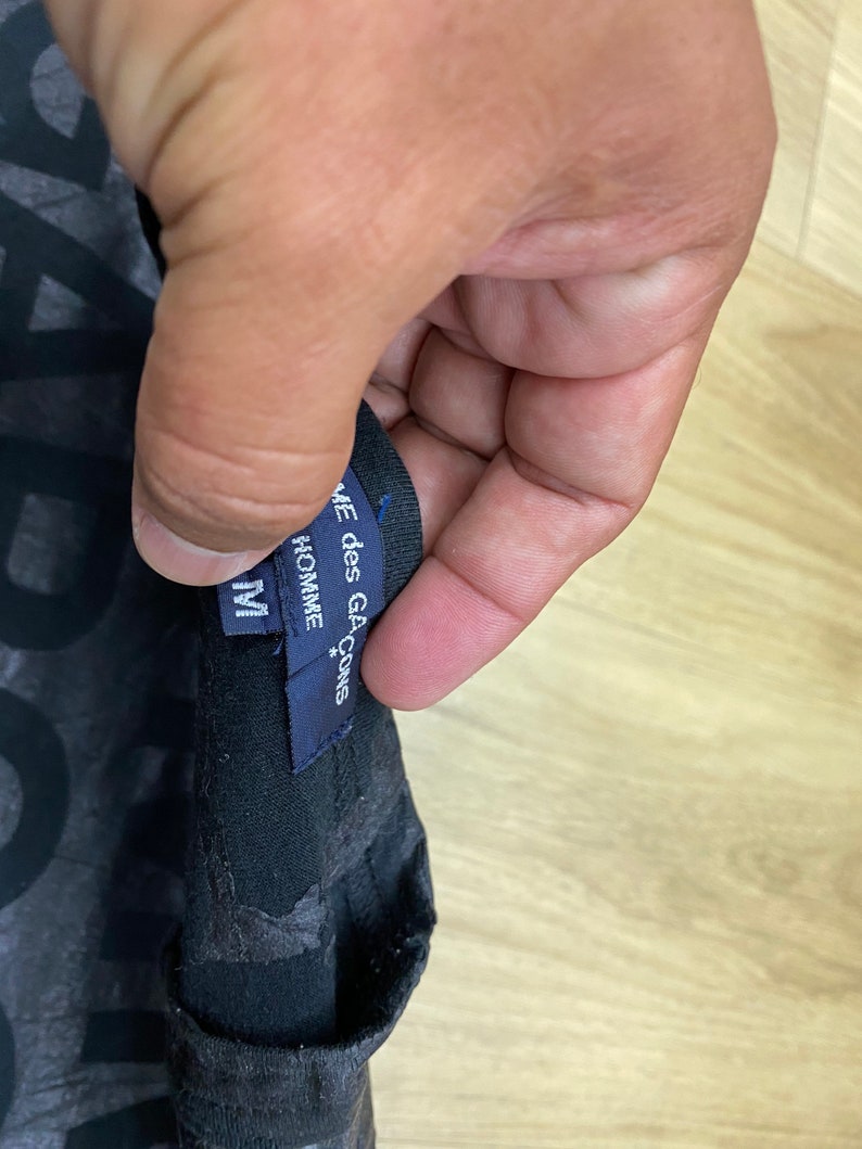 May include: A black t-shirt with a graphic print and a blue tag that reads "COMME des GARCONS*" and "HOMME" with a size "M" label.