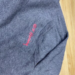 May include: A dark gray long-sleeved shirt with a pocket. The text "OK-Hang" is embroidered in pink on the upper left side. The shirt is folded on a light wood surface.