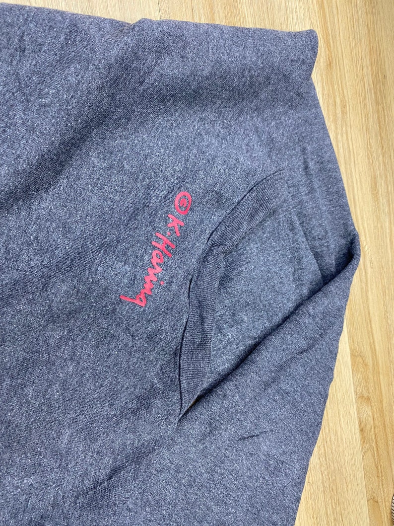 May include: A dark gray long-sleeved shirt with a pocket. The text "OK-Hang" is embroidered in pink on the upper left side. The shirt is folded on a light wood surface.