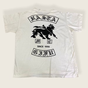 May include: White t-shirt with a black graphic of a lion with a crown, a spear, and the text "RASTA" and "G.I.F.U." in a gothic font. The text "SINCE 1999" is also printed on the shirt.