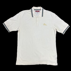 Puede incluir: Polo blanco con cuello a rayas blanco y negro y un logotipo bordado en dorado en el pecho.