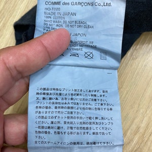 May include: A white tag with black text and symbols. The tag is attached to a black garment. The text on the tag reads "COMME des GAR&Ccedil;ONS Co., Ltd. HO-T020 MADE IN JAPAN 100% COTTON HAND WASH. DO NOT BLEACH. DO NOT IRON. DO NOT DRY CLEAN. LINE DRY FAR I JAPON EL INTERDIT. NE FERDIT NETTOYAGE A L'OMBRE." The tag also includes symbols for hand washing, no bleaching, no ironing, no dry cleaning, and line drying.
