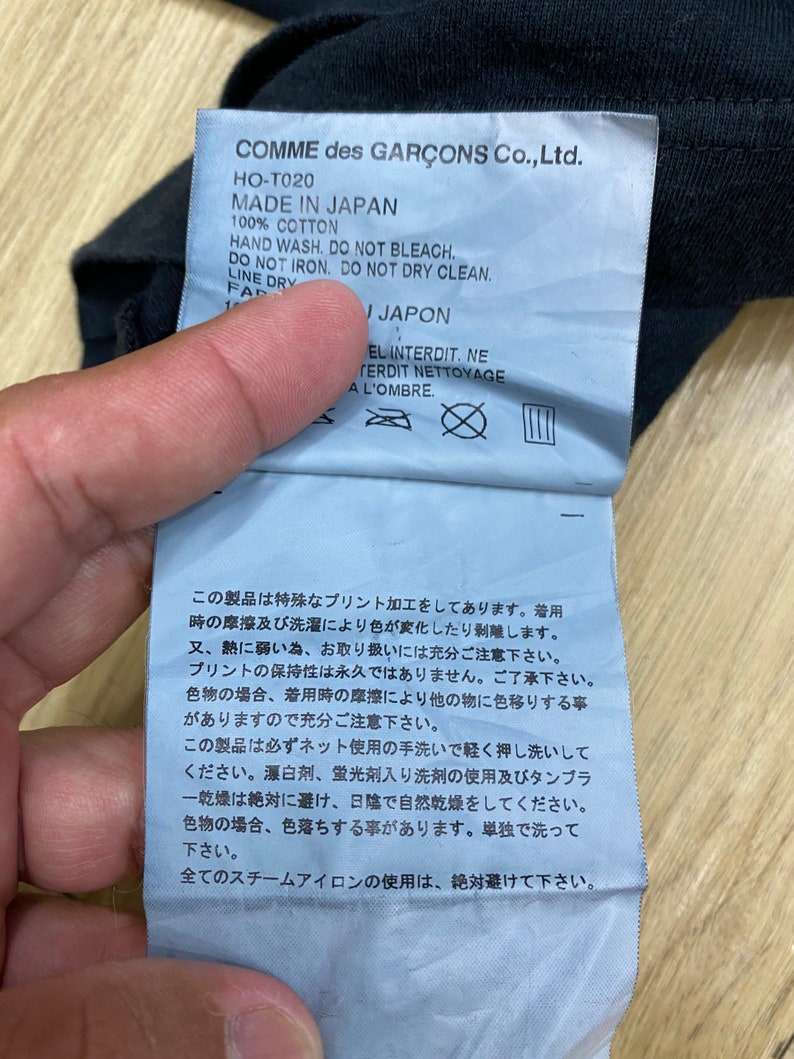 May include: A white tag with black text and symbols. The tag is attached to a black garment. The text on the tag reads "COMME des GAR&Ccedil;ONS Co., Ltd. HO-T020 MADE IN JAPAN 100% COTTON HAND WASH. DO NOT BLEACH. DO NOT IRON. DO NOT DRY CLEAN. LINE DRY FAR I JAPON EL INTERDIT. NE FERDIT NETTOYAGE A L'OMBRE." The tag also includes symbols for hand washing, no bleaching, no ironing, no dry cleaning, and line drying.
