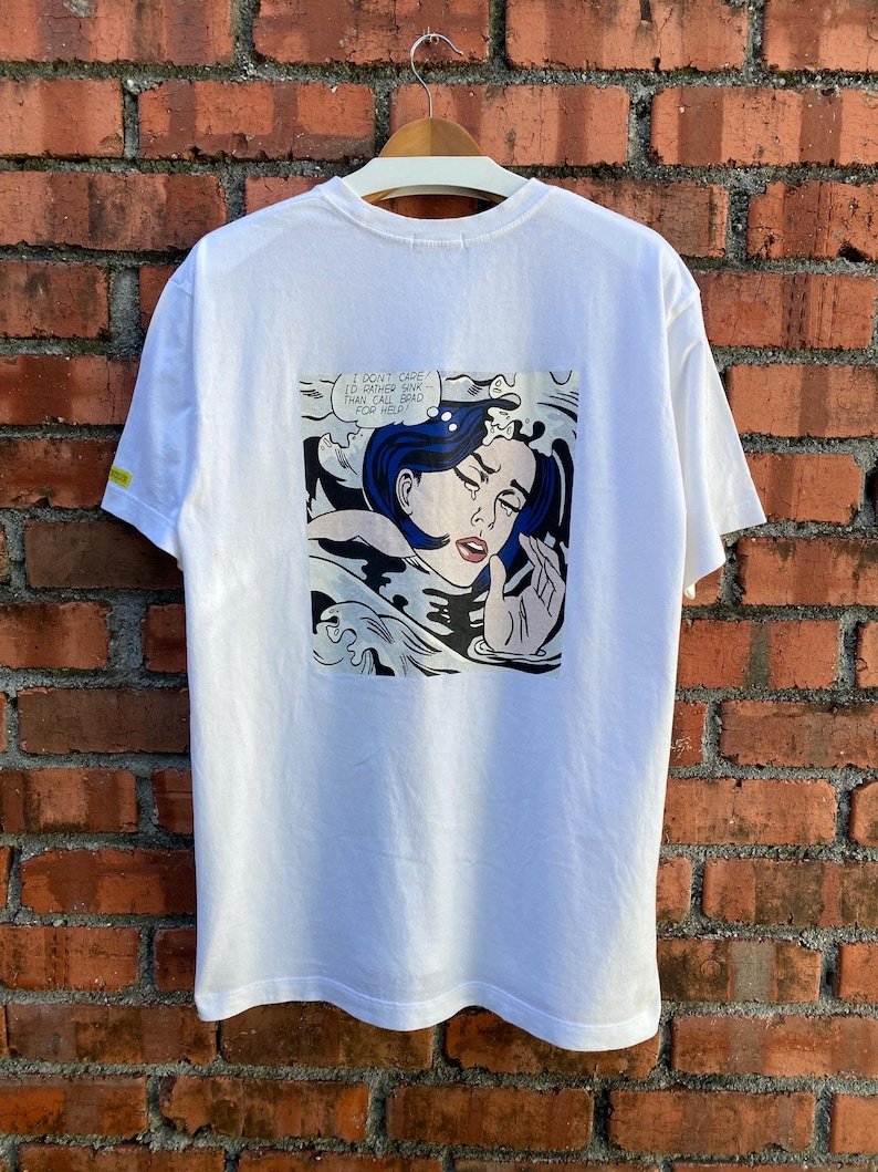 May include: White t-shirt featuring a pop art comic strip on the back. The artwork depicts a crying woman with blue hair in a body of water, with the text "I DON'T CARE! I'D RATHER SINK THAN CALL BRAD FOR HELP!"