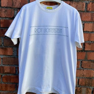 May include: White t-shirt with the text "ROY LICHTENSTEIN" in gray, above and below the text are rows of gray dots. The shirt is hanging on a wooden hanger against a brick wall.