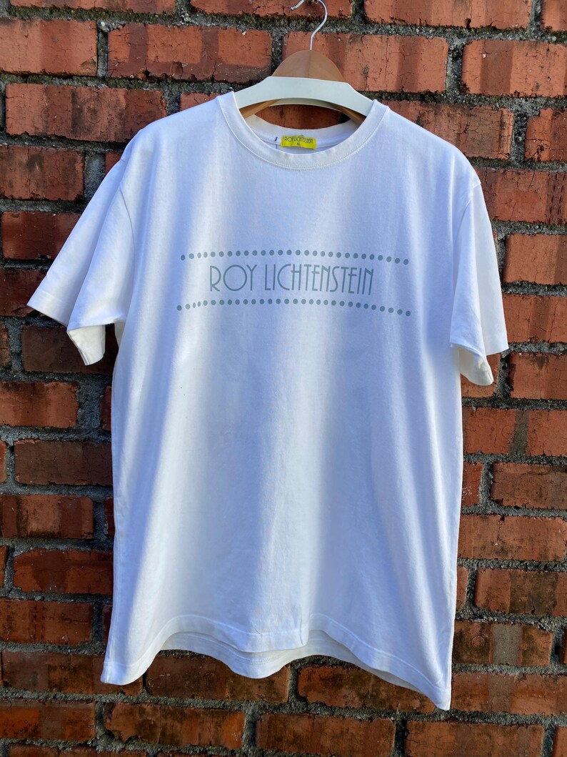 May include: White t-shirt with the text "ROY LICHTENSTEIN" in gray, above and below the text are rows of gray dots. The shirt is hanging on a wooden hanger against a brick wall.