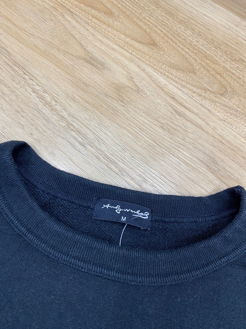 Puede incluir: Sudadera negra con una etiqueta negra que dice "And Wander" y "M" para el tama&ntilde;o.